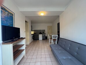 A living room with a grey sofa, a flat-screen TV on a stand, and a kitchen in the background. - Ground floor apart, walking distance to the beach (Cabourg)