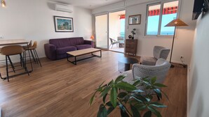 A modern living room with a sofa, coffee table, and chairs. There is a dining area with a table and chairs. The room has wooden flooring and a large window with a view. - Sea view apartment, Sanguinaires Islands terraces (Ajaccio)
