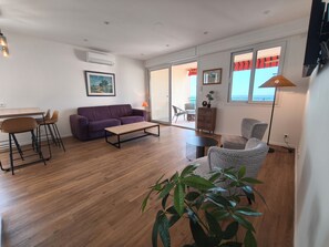 Ein modernes Wohnzimmer mit Sofa, Couchtisch und Sesseln. Es gibt einen Essbereich mit Tisch und Stühlen. Der Raum hat Holzböden und ein großes Fenster mit Blick.