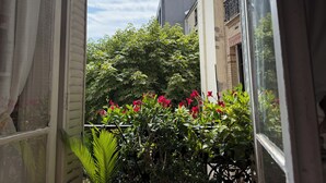 Blick durch ein Fenster mit Balkon, zeigt ein Gebäude, Grünflächen und einen klaren Himmel.