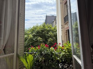 A view through a window with a balcony, featuring a building, greenery, and a clear sky. - Large, quiet apartment with balcony in Paris 12 (Paris)