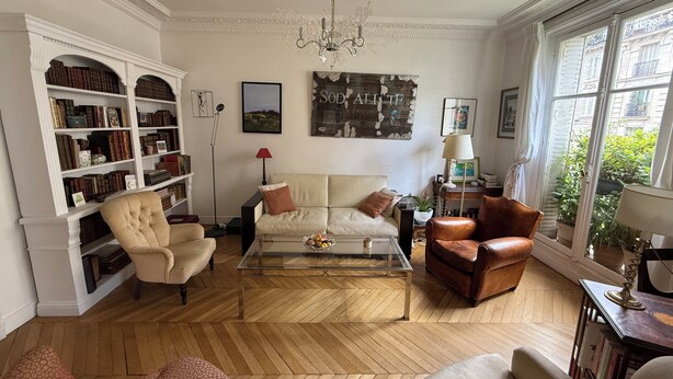 Ein geräumiges Wohnzimmer mit einer großen Bücherwand, einer Sofagarnitur, einem Couchtisch und Blick ins Freie durch ein Fenster.