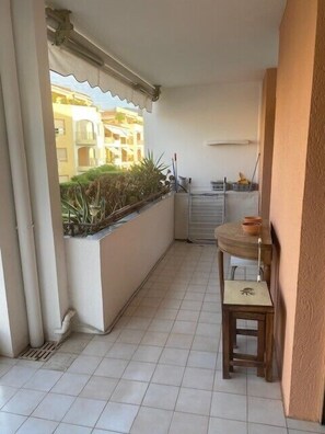 Un balcó estret amb un paviment de rajoles, un tamboret de fusta i una finestra amb vista a una zona residencial.