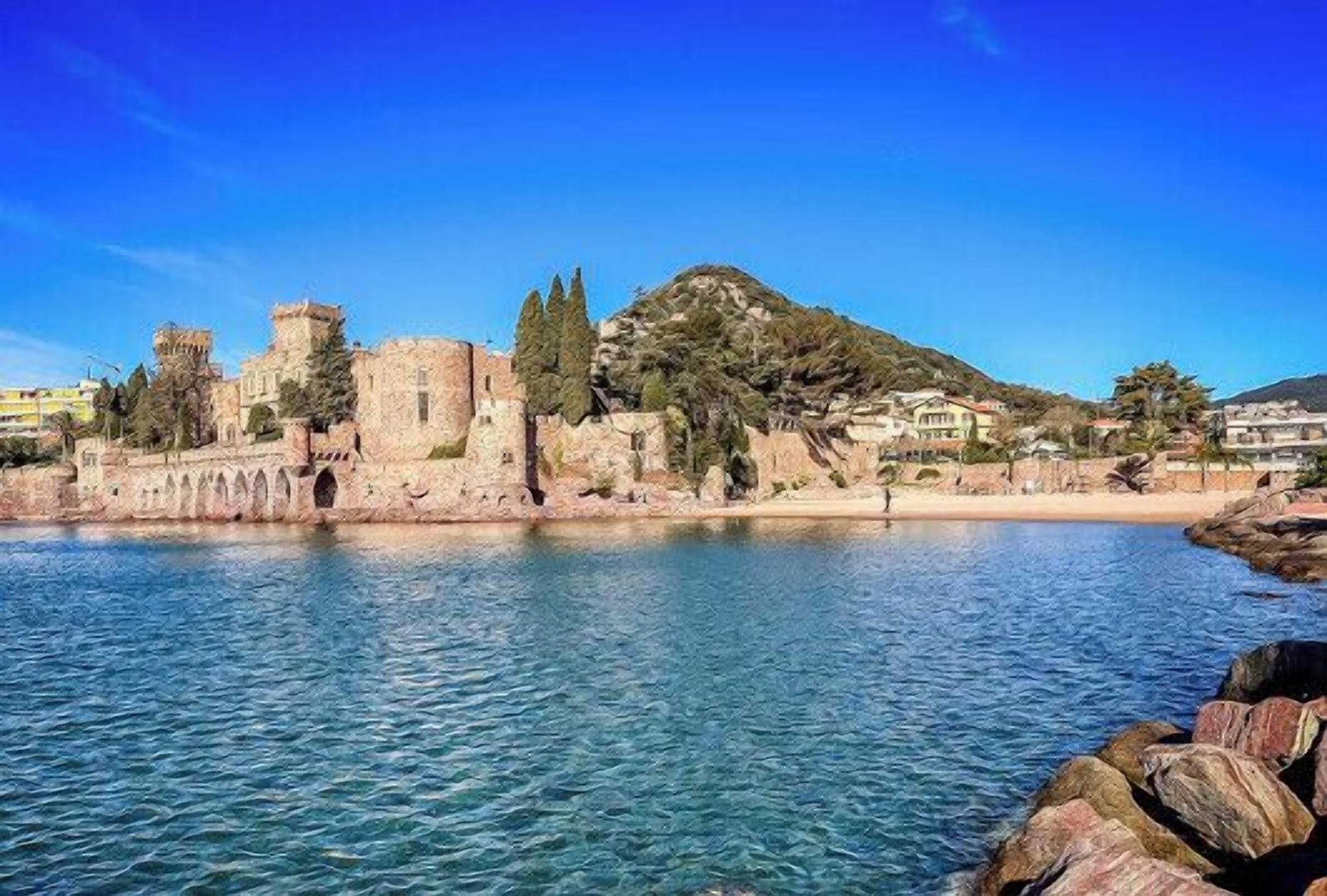 A coastal cityscape with a historic castle, clear blue waters, and a rocky shoreline.