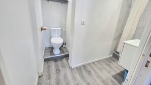 Een badkamer met toilet, douche en een plank met een vaas en bekers.