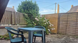 Een terras met een tafel en twee stoelen, omgeven door een houten schutting en een struik met roze bloemen.