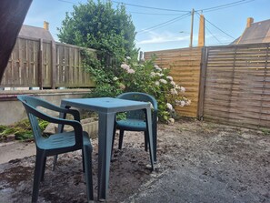 Eine Terrasse mit Tisch und zwei Stühlen, umgeben von einem Holzzaun und einem Busch mit rosafarbenen Blüten.