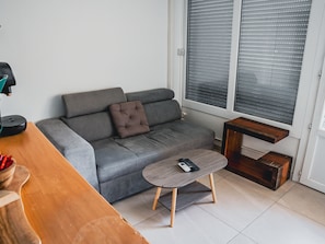 Ein modernes Wohnzimmer mit einer grauen Couch, einem kleinen Holztisch und einem Fenster mit Jalousien.