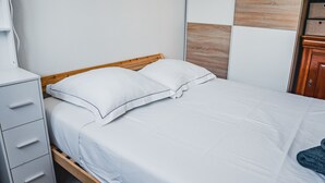 En pænt redt seng med hvide sengetøj og to puder, en træsenggavl, et hvidt natbord med skuffer, og en træskab.