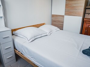 Ein ordentlich bezogenes Bett mit weißen Bettwäsche und zwei Kissen, ein Holzkopfstück, ein weißer Nachttisch mit Schubladen und ein Holzschrank.