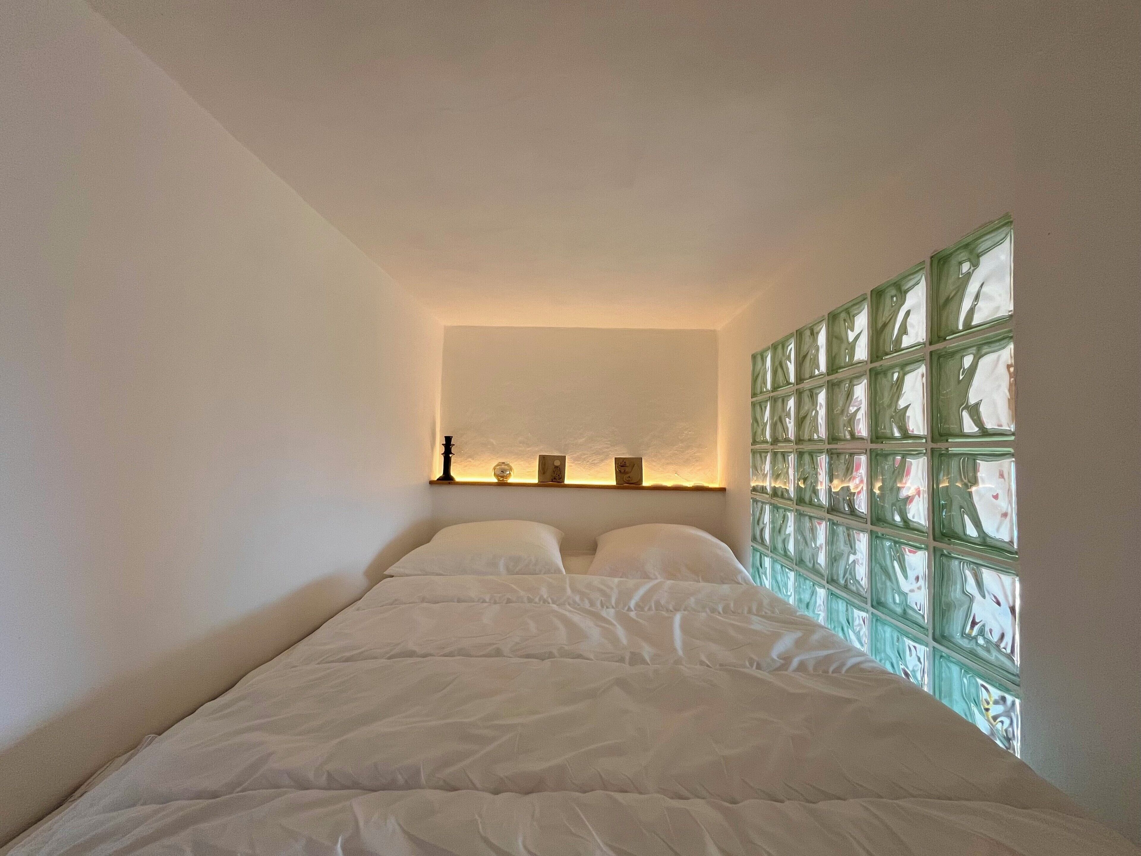 Um quarto com uma cama, uma prateleira com objetos decorativos e uma janela com blocos de vidro verde.
