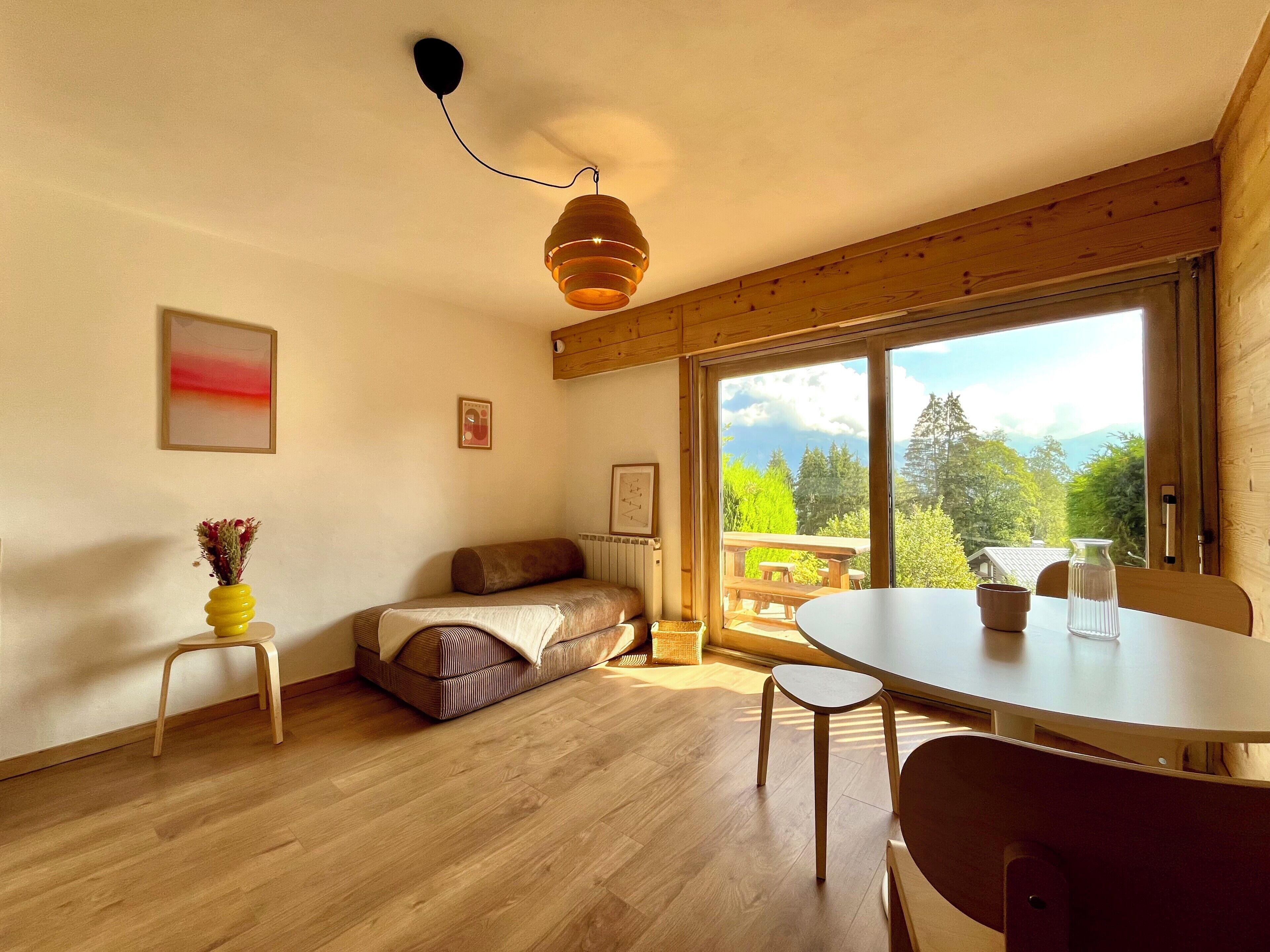 Une pièce chaleureuse avec un plancher en bois, un canapé, une table à manger avec des chaises, un luminaire suspendu et une grande fenêtre donnant sur un paysage verdoyant et montagneux.