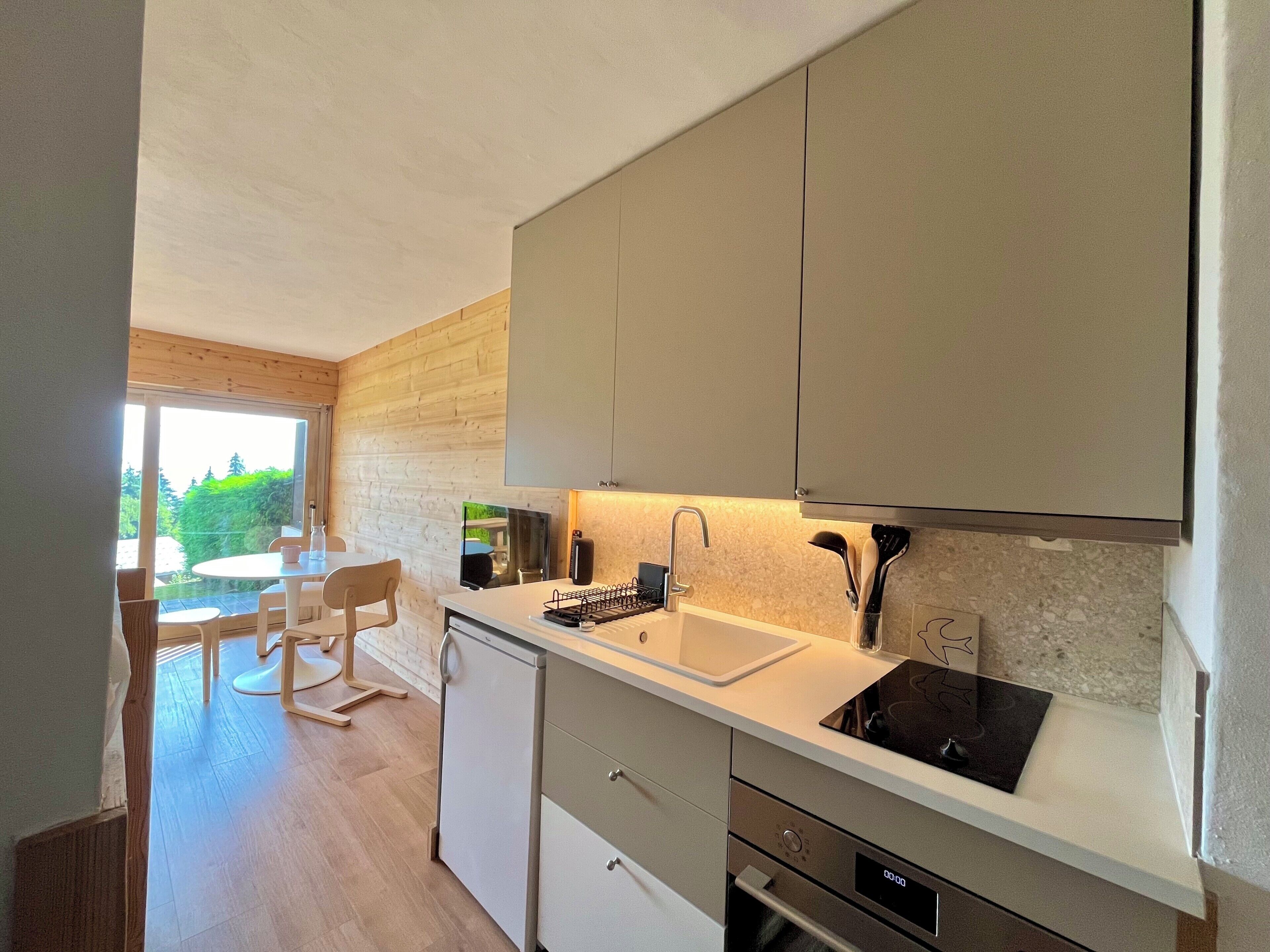 A modern kitchen with a dishwasher, oven, and a sink with a faucet. There is a dining area with a table and chairs visible through a glass door.