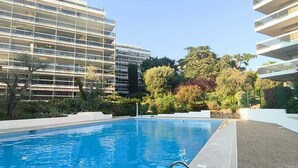 Una piscina con escalera metálica, rodeada de un área ajardinada con árboles y arbustos, y contigua a edificios modernos de varios pisos.