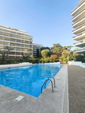A swimming pool with a metal ladder, surrounded by a landscaped area with trees and shrubs, and adjacent to modern multi-story buildings. - Apartment with sea view, terrace and swimming pool (Antibes)