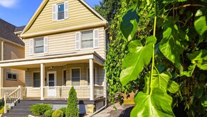 Une maison à deux étages avec un porche à l’avant, un petit jardin devant et un mur recouvert de plantes grimpantes.