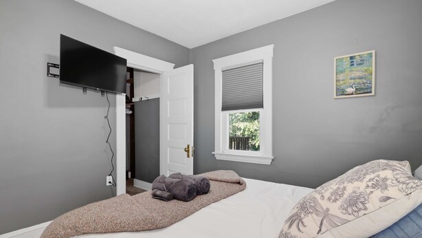 Une chambre à coucher avec un lit, un téléviseur fixé au mur, une fenêtre avec des stores et un tableau accroché au mur.