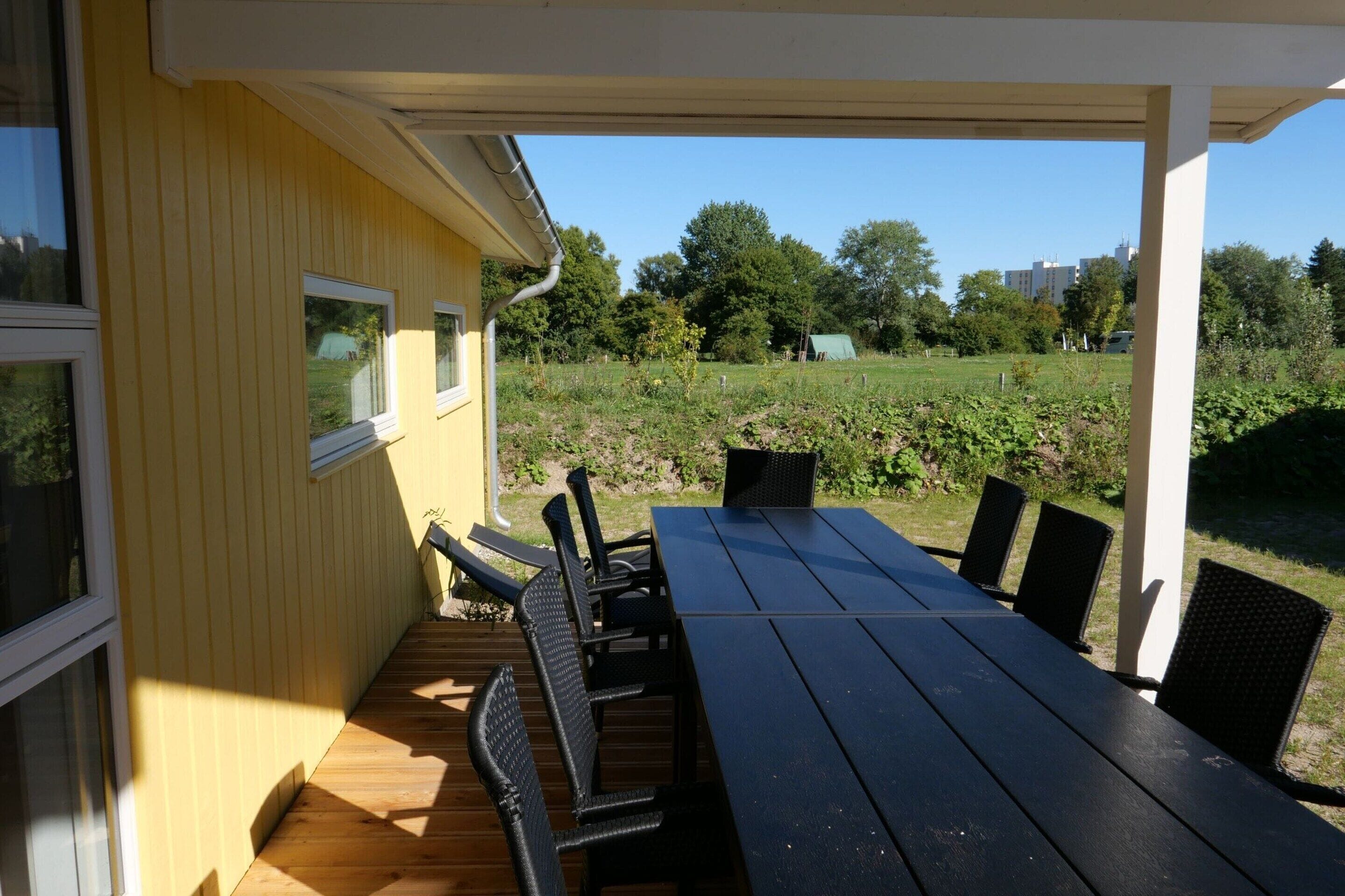 Un espacio al aire libre cubierto con una mesa y sillas, una terraza de madera y vistas a un campo verde y árboles.