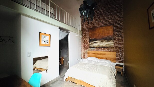 Une chambre avec un lit en mezzanine, un bureau, une chaise, un luminaire suspendu et un mur en briques.