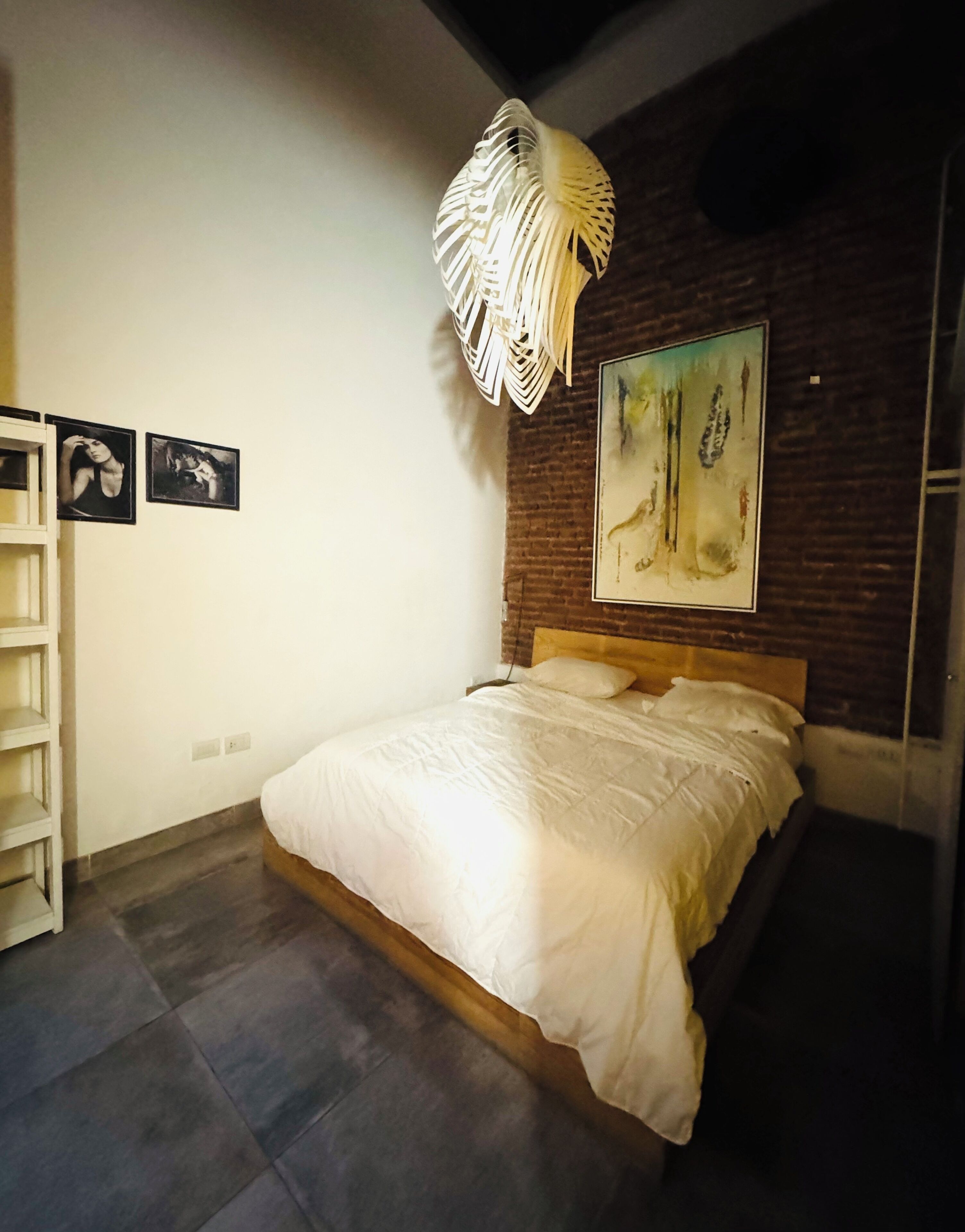 A bedroom with a bed, a white headboard, a wooden floor, a brick wall, and a hanging light fixture.
