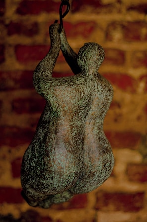 A bronze sculpture of a human figure with outstretched arms, hanging against a brick wall background.