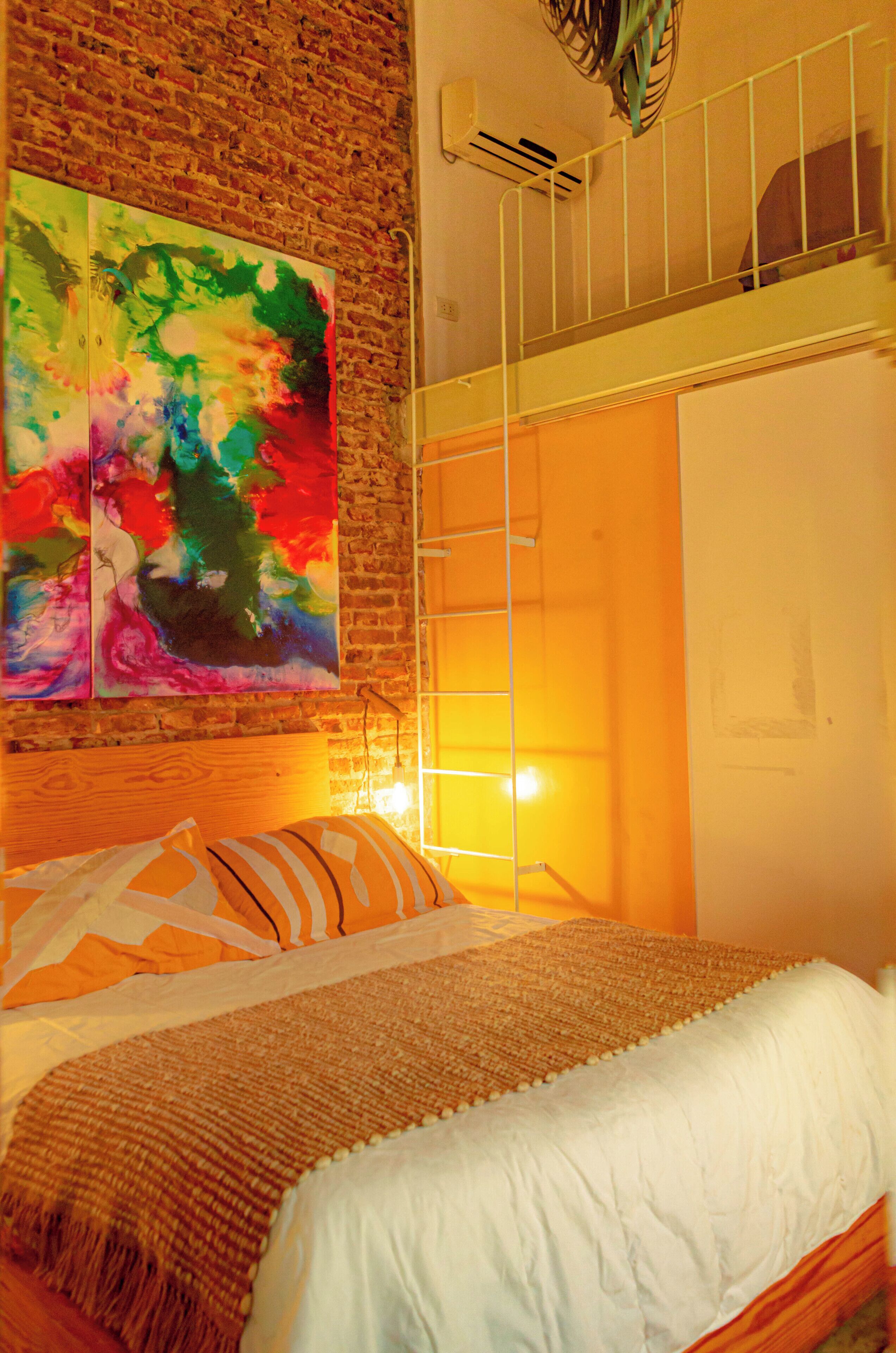 Un dormitorio con pared de ladrillo, cama tipo loft, un cuadro abstracto de colores y una lámpara de noche.