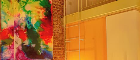 A bedroom with a brick wall, a loft bed, a colorful abstract painting, and a bedside lamp.