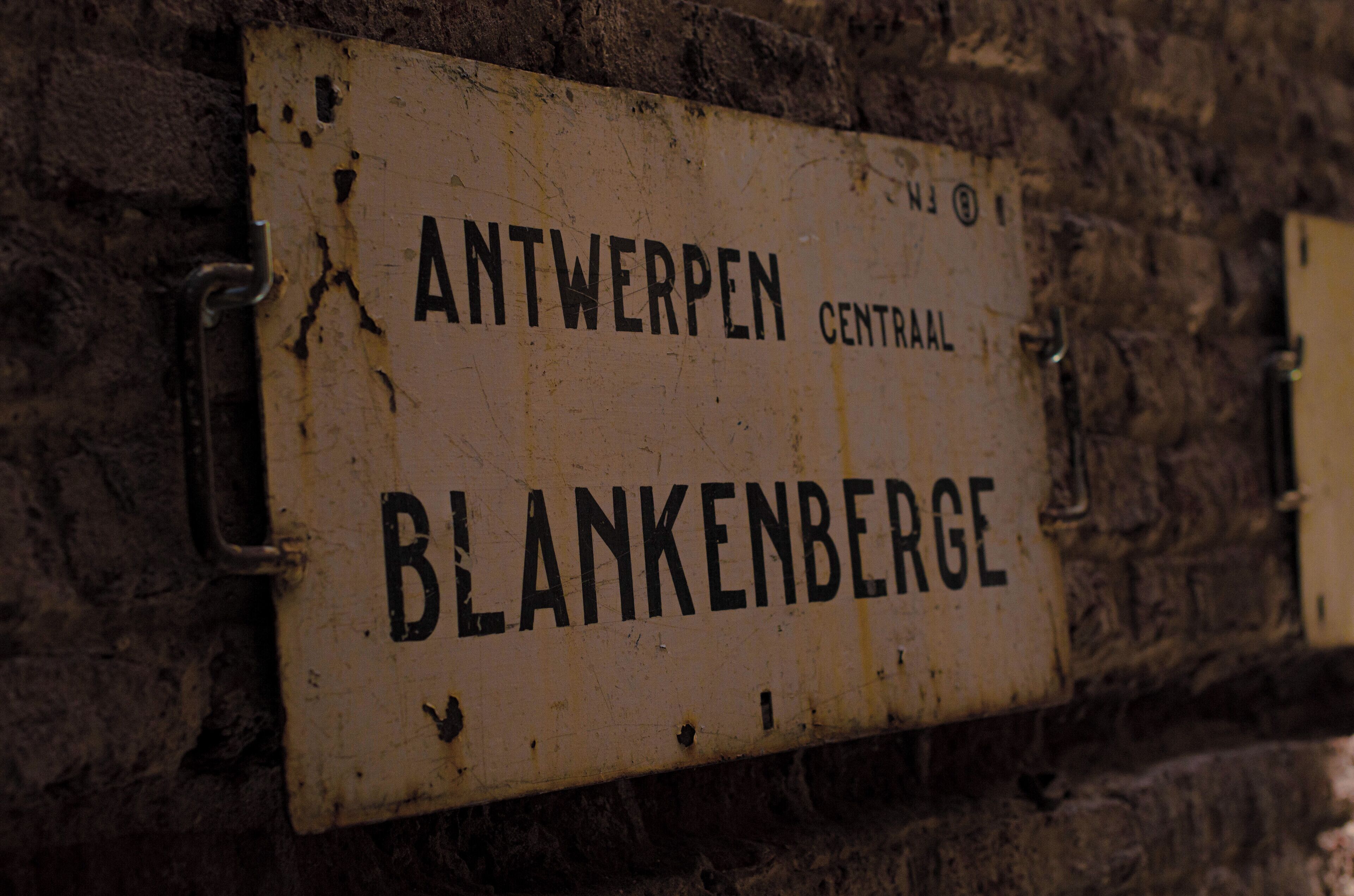 Un letrero desgastado con la inscripción 'ANTWERPEN CENTRAL BLANKENBERGE' sobre un muro de ladrillo.