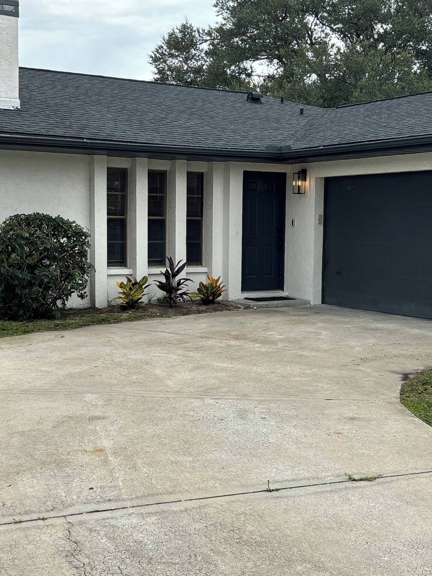 A single-story house with a dark-colored garage door, a front entrance with a black door, and a small front yard with a bush and a few potted plants.