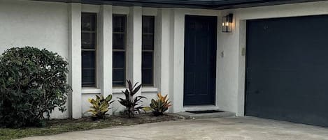 A single-story house with a dark-colored garage door, a front entrance with a black door, and a small front yard with a bush and a few potted plants.