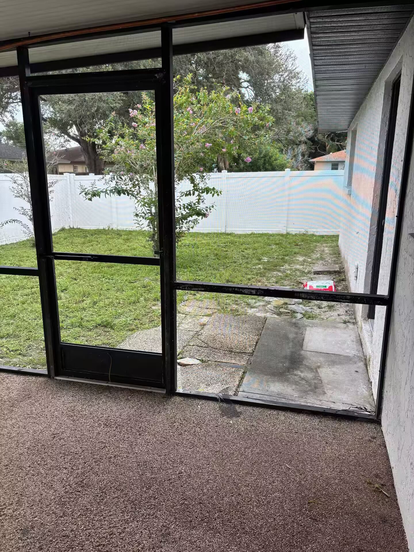 A view through a sliding glass door into a backyard with a grassy area, a concrete patio, and a white fence.