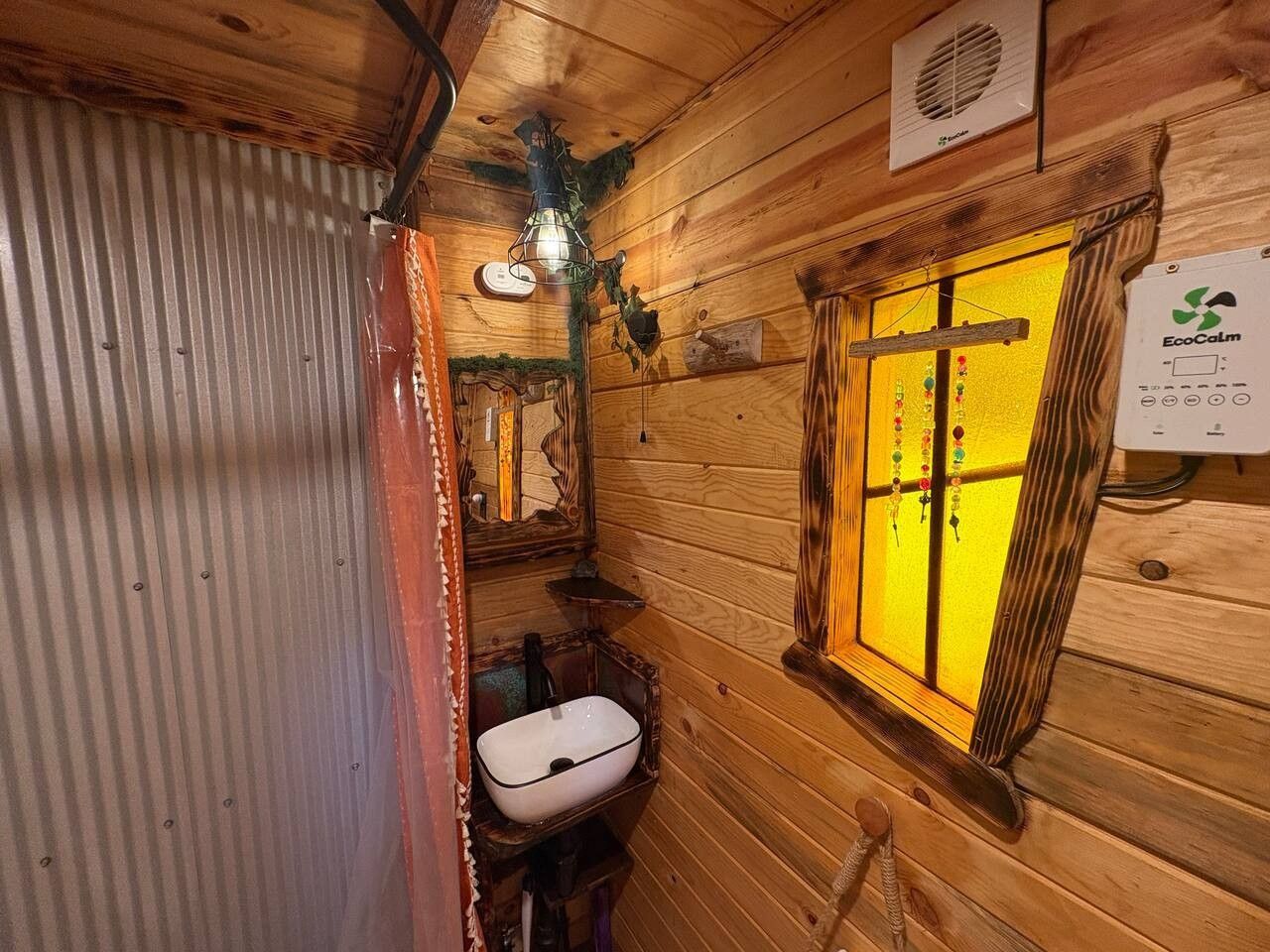 A wooden cabin interior with a wooden bench, a white sink, a window with curtains, and a wall-mounted EcoCalm device.