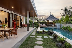 A modern outdoor pool area with a stone pathway leading to a gazebo, surrounded by lush greenery and palm trees.