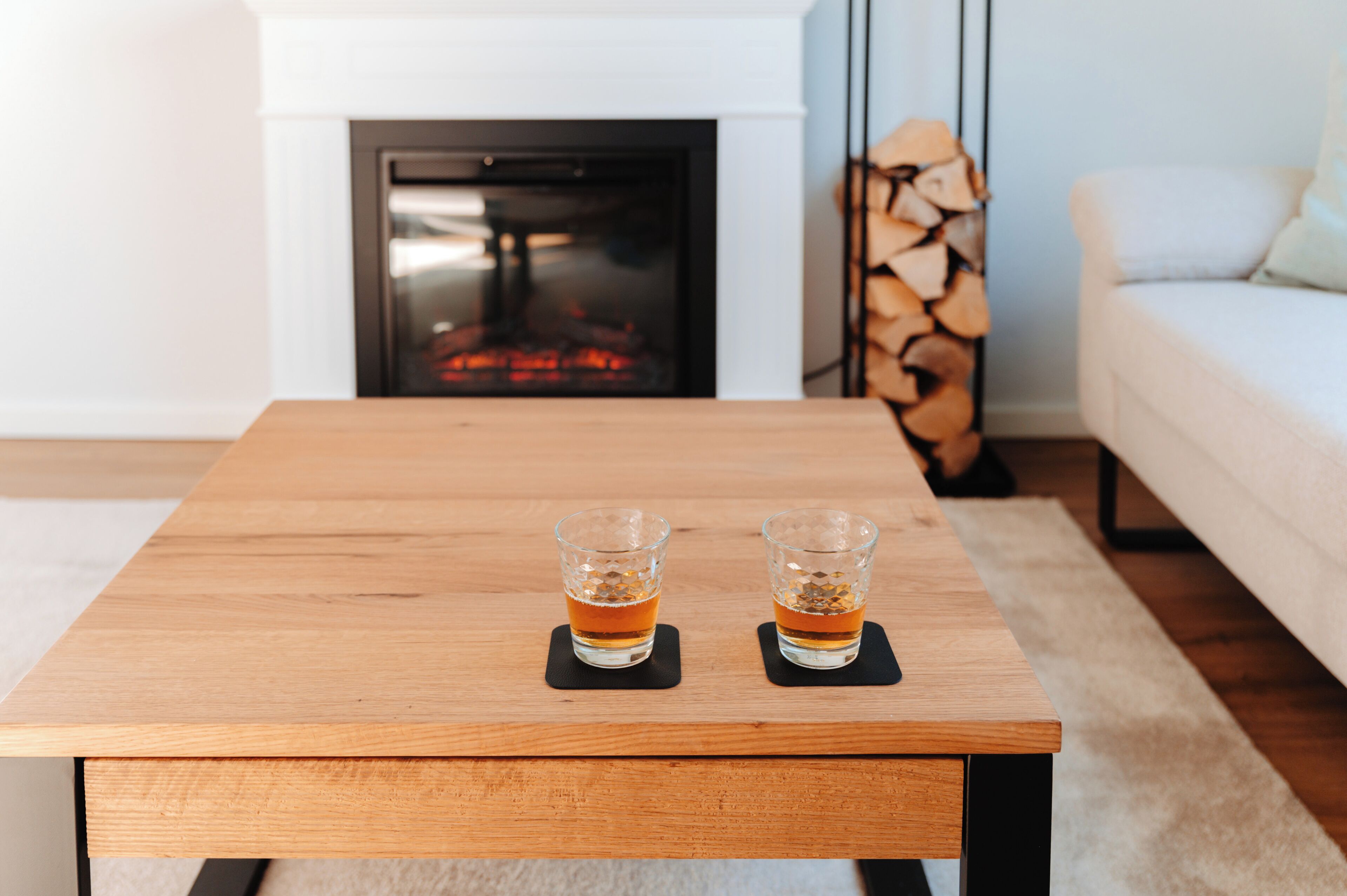 Ein Holztisch mit zwei Gläsern bernsteinfarbener Flüssigkeit, vermutlich Whiskey, auf schwarzen Untersetzern.