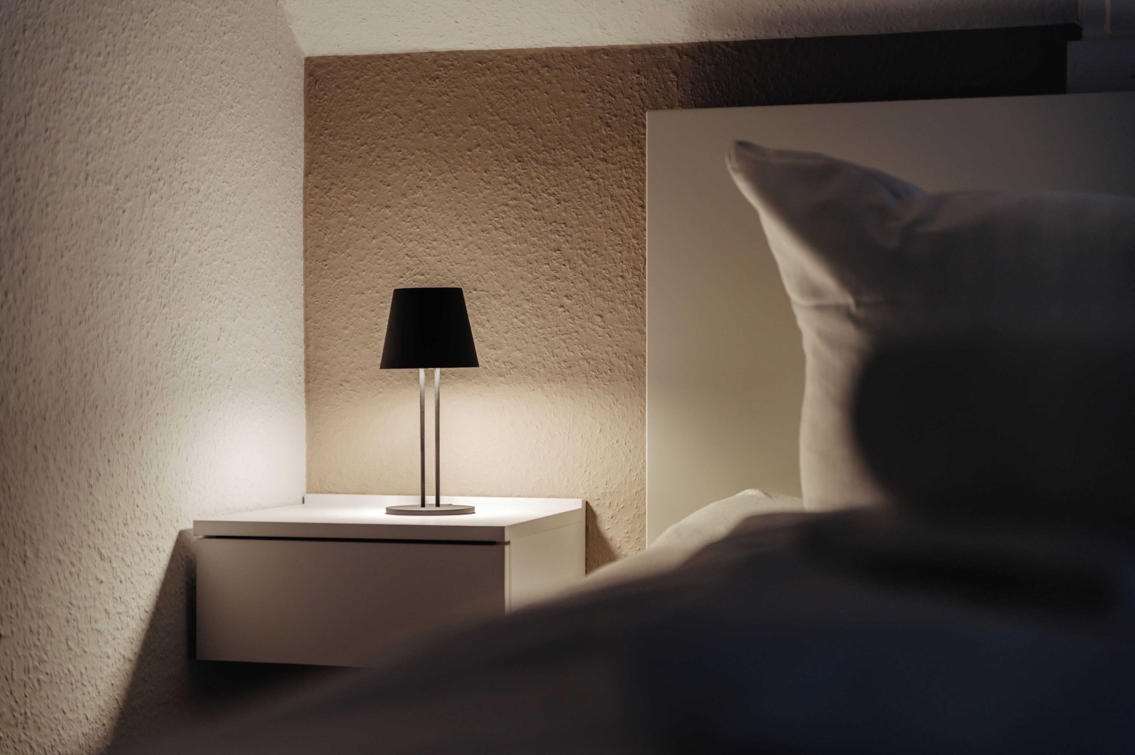 Ein Nachttisch mit Lampe, eine Wand mit strukturierter Oberfläche und ein weißes Kissen.