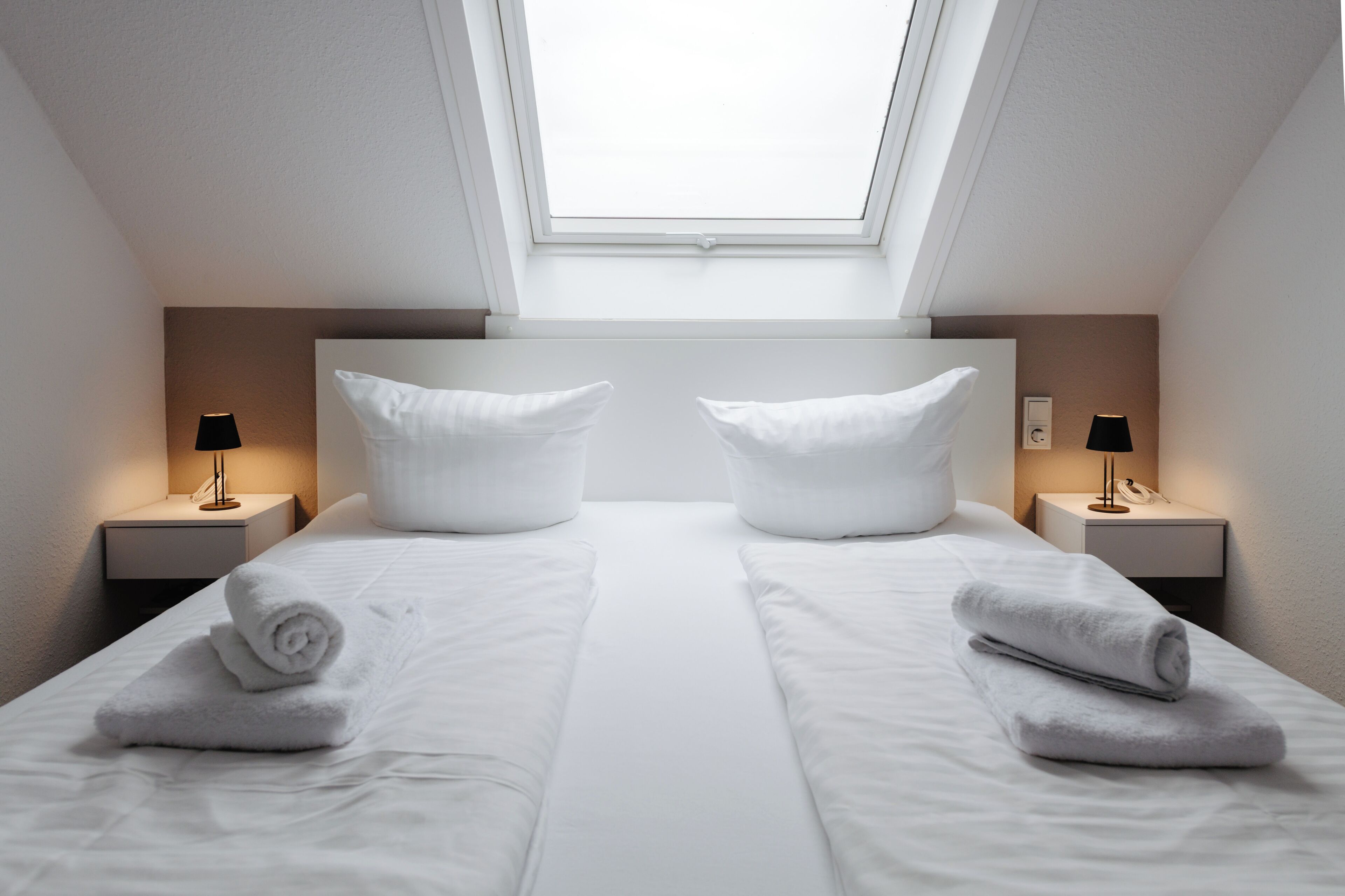 Ein Doppelzimmer mit Oberlichter, zwei Nachttische inklusive Lampen und ordentlich gefaltete Handtücher.