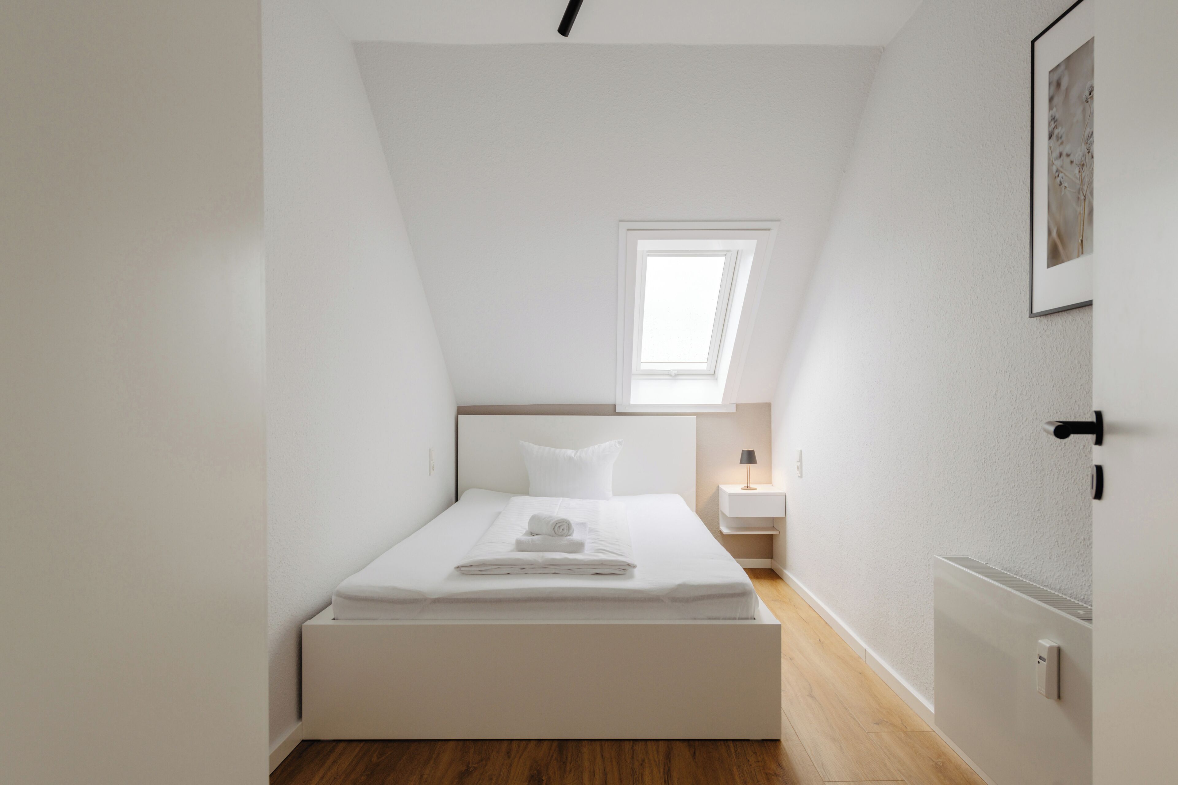 A minimalist bedroom with a single bed, a bedside table, a nightstand, a window, and a radiator.