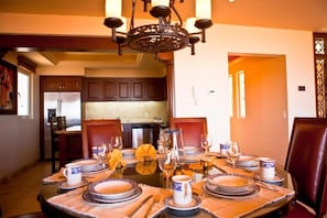 A dining area with a round table set for six, surrounded by chairs. A chandelier hangs above the table, and the room has wooden cabinets and a kitchen in the background. - Pueblo Bonito Montecristo Luxury Villas - All Inclusive (Cabo San Lucas)