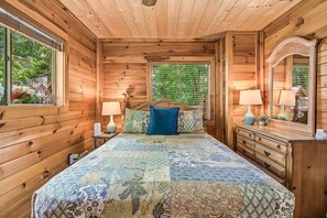 A wooden cabin bedroom with a bed, bedside lamps, a dresser, and a window with a view of greenery. - Halfway to Heaven | Hot Tub, Game Room & Views (Gatlinburg)