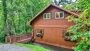 A wooden cabin with a porch, surrounded by dense forest. - Halfway to Heaven | Hot Tub, Game Room & Views (Gatlinburg)