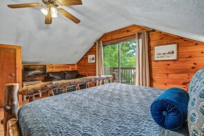 A bedroom with a sloped ceiling, a bed with a patterned bedspread, a wooden headboard, a ceiling fan, and a window with curtains. - Halfway to Heaven | Hot Tub, Game Room & Views (Gatlinburg)