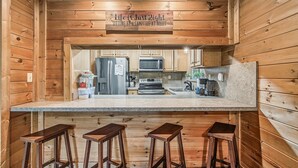 A kitchen with wooden walls, a granite countertop, stainless steel appliances, and three wooden stools. - Halfway to Heaven | Hot Tub, Game Room & Views (Gatlinburg)