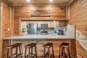 A kitchen with wooden walls, a granite countertop, stainless steel appliances, and three wooden stools. - Halfway to Heaven | Hot Tub, Game Room & Views (Gatlinburg)