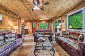A log cabin interior with a central seating area, wooden walls, and a large window providing a view of the outdoors. - Halfway to Heaven | Hot Tub, Game Room & Views (Gatlinburg)