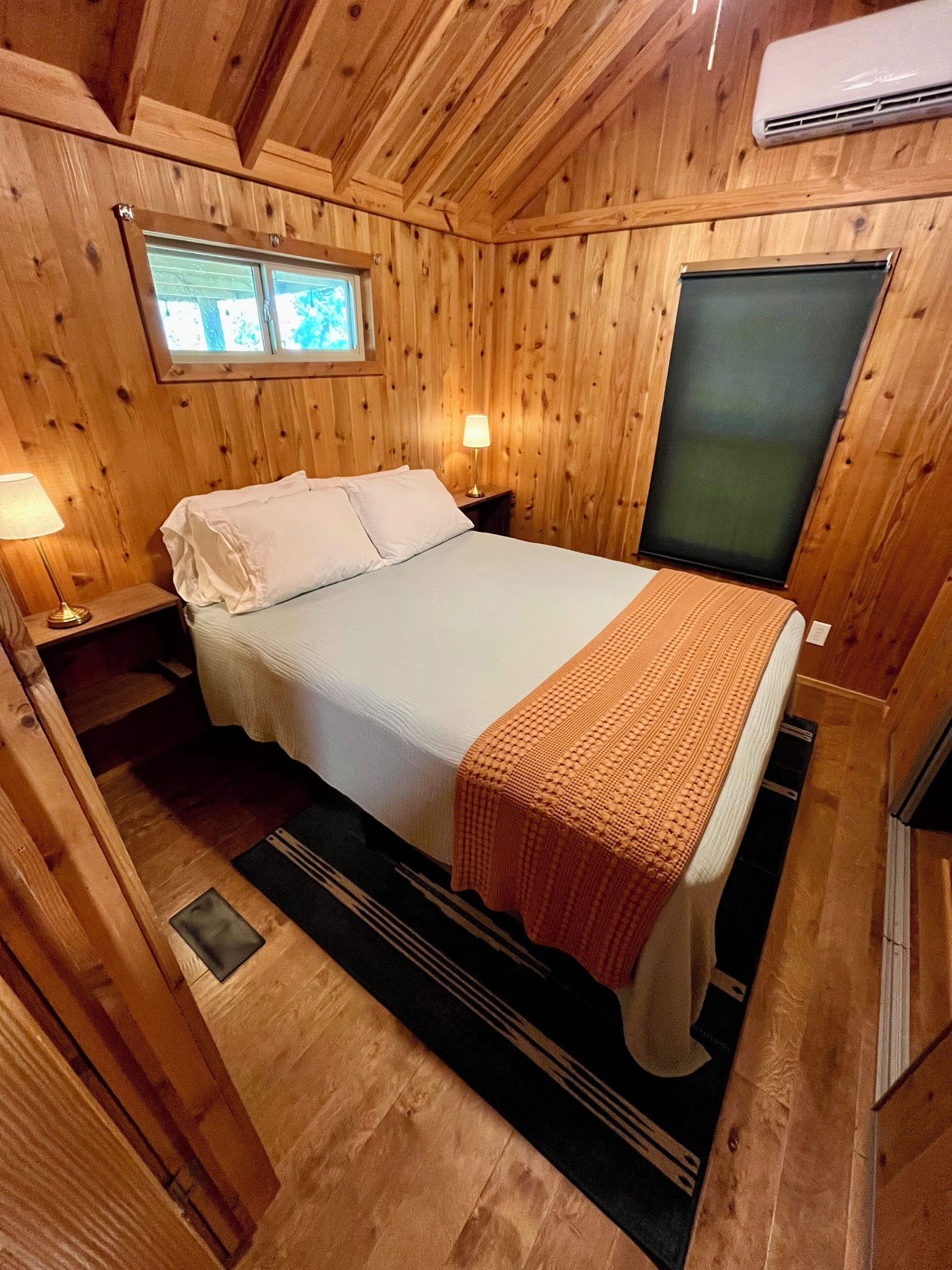 Une chambre dans une cabane en bois, comprenant un lit, des tables de chevet, une fenêtre et un appareil de climatisation.