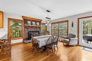 Une salle à manger avec une cheminée en pierre, des chaises en bois, une table recouverte d’une nappe blanche et de grandes fenêtres donnant sur la forêt.