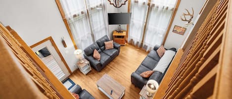 A spacious living room with a modern chandelier, wooden flooring, and a large window with sheer curtains.