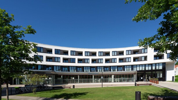 En modern byggnad med flera våningar, en böjd fasad, stora fönster och en central innergård med en gräsyta och bänkar.