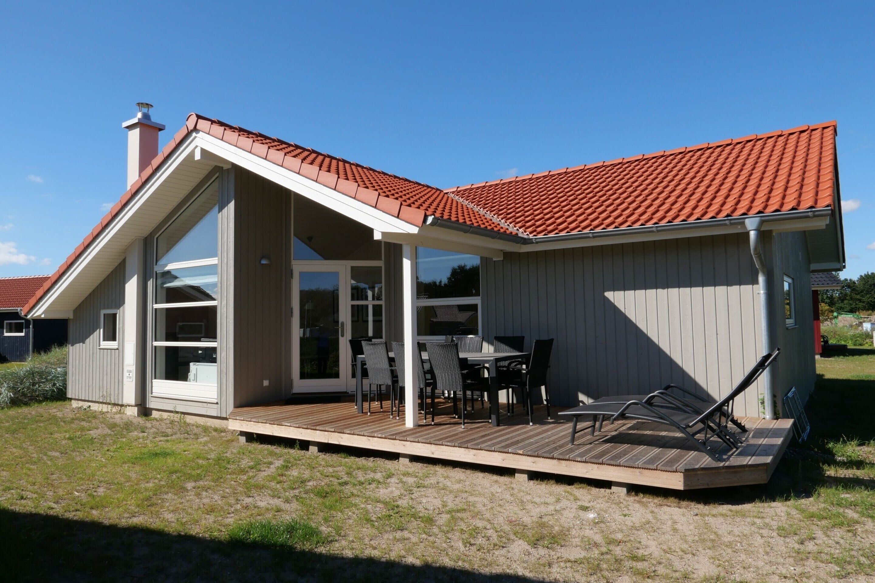Une maison moderne avec un toit recouvert de tuiles rouges, un patio couvert avec des meubles d’extérieur et une grande porte vitrée donnant sur l’intérieur.