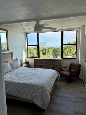 A bedroom with a bed, a desk, a chair, and a view of greenery through large windows.